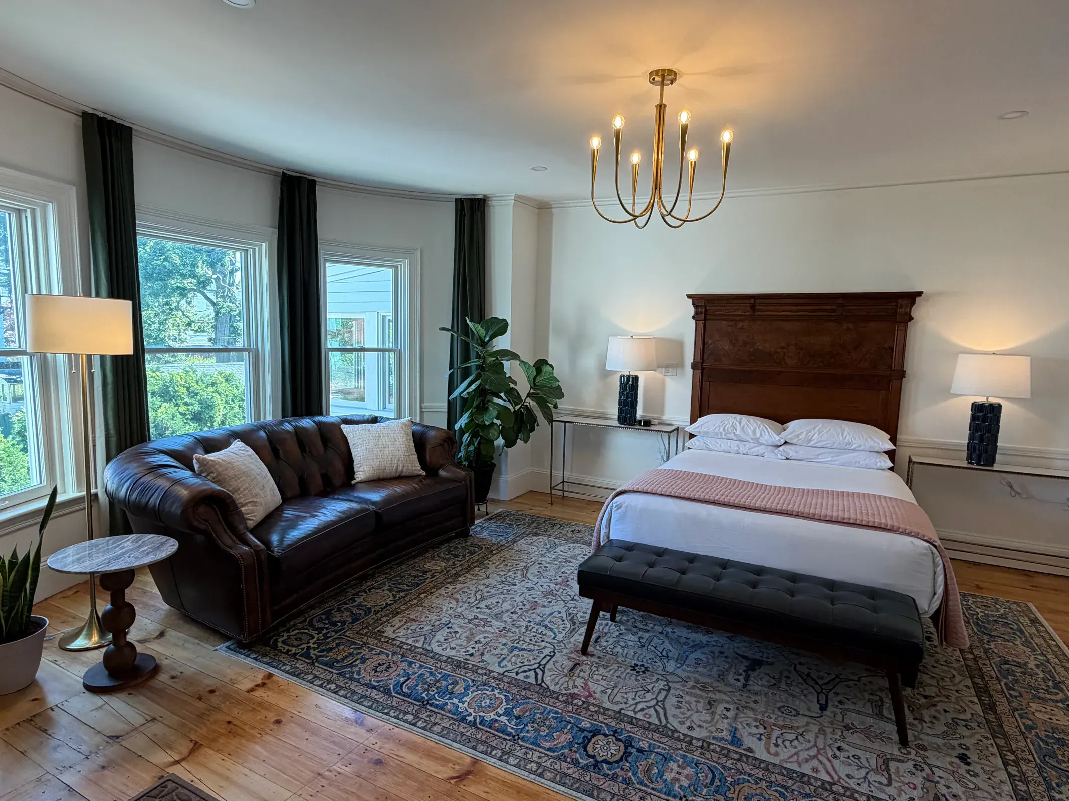 A bright Delphian guest room with a bed, leather sofa, hardwood floors, and tall windows